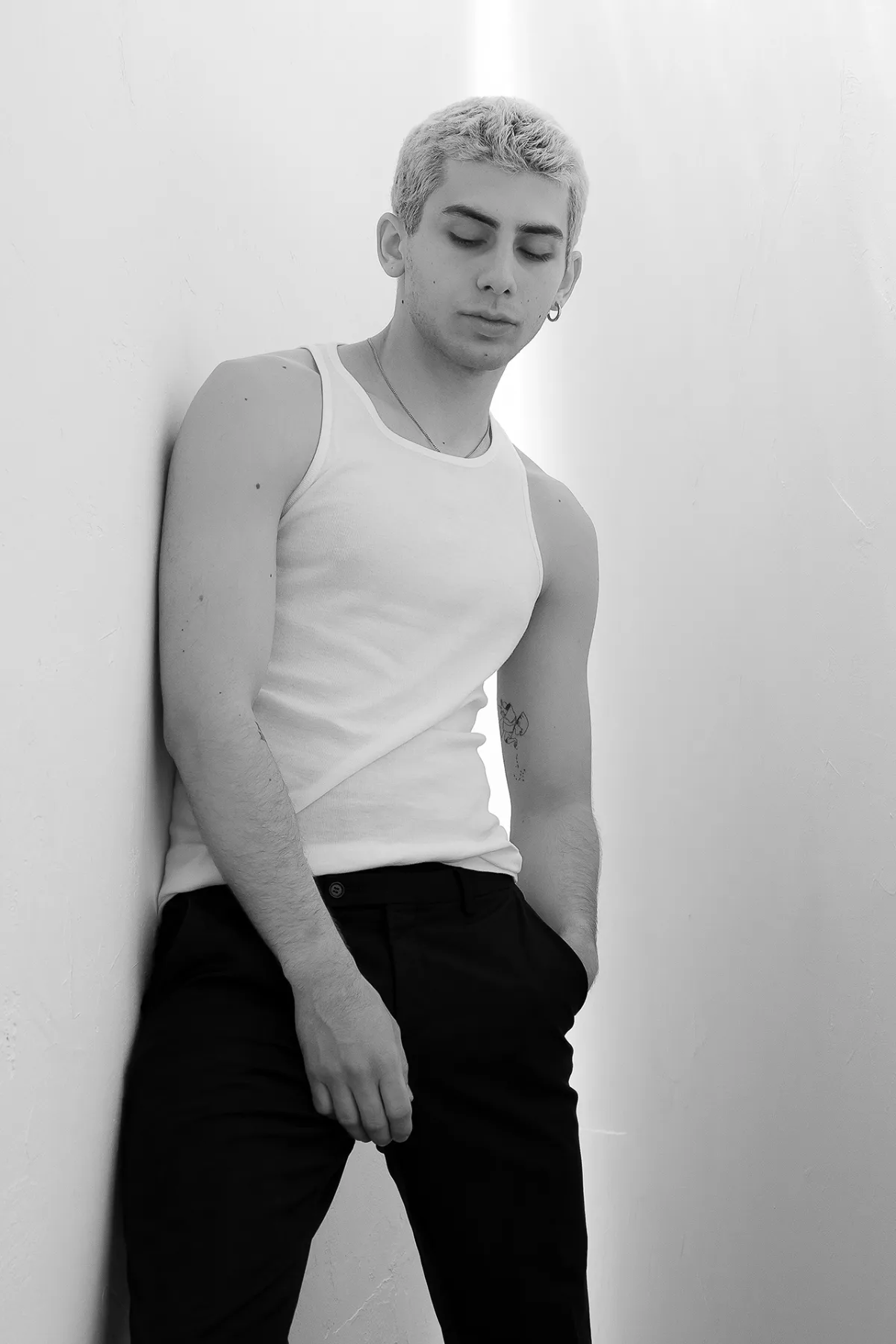 Black-and-white beauty portrait of a young man with light hair, white tank top, leaning against a wall