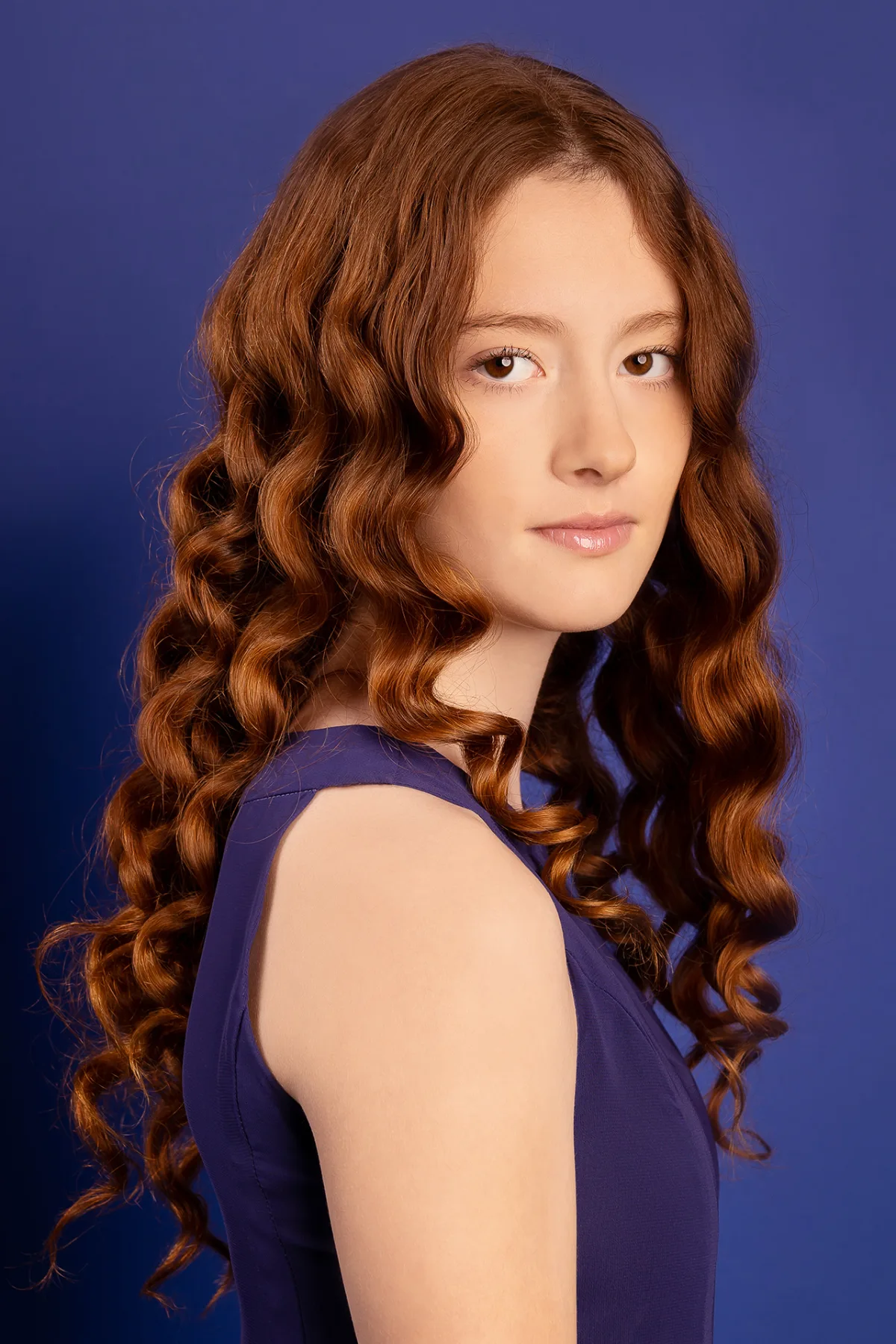 Beauty portrait of a girl with long red curls, blue studio backdrop, blue dress
