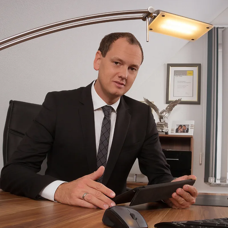 Business portrait: a man in a dark suit and grey tie at his desk, holding a tablet, with a desk lamp in the foreground