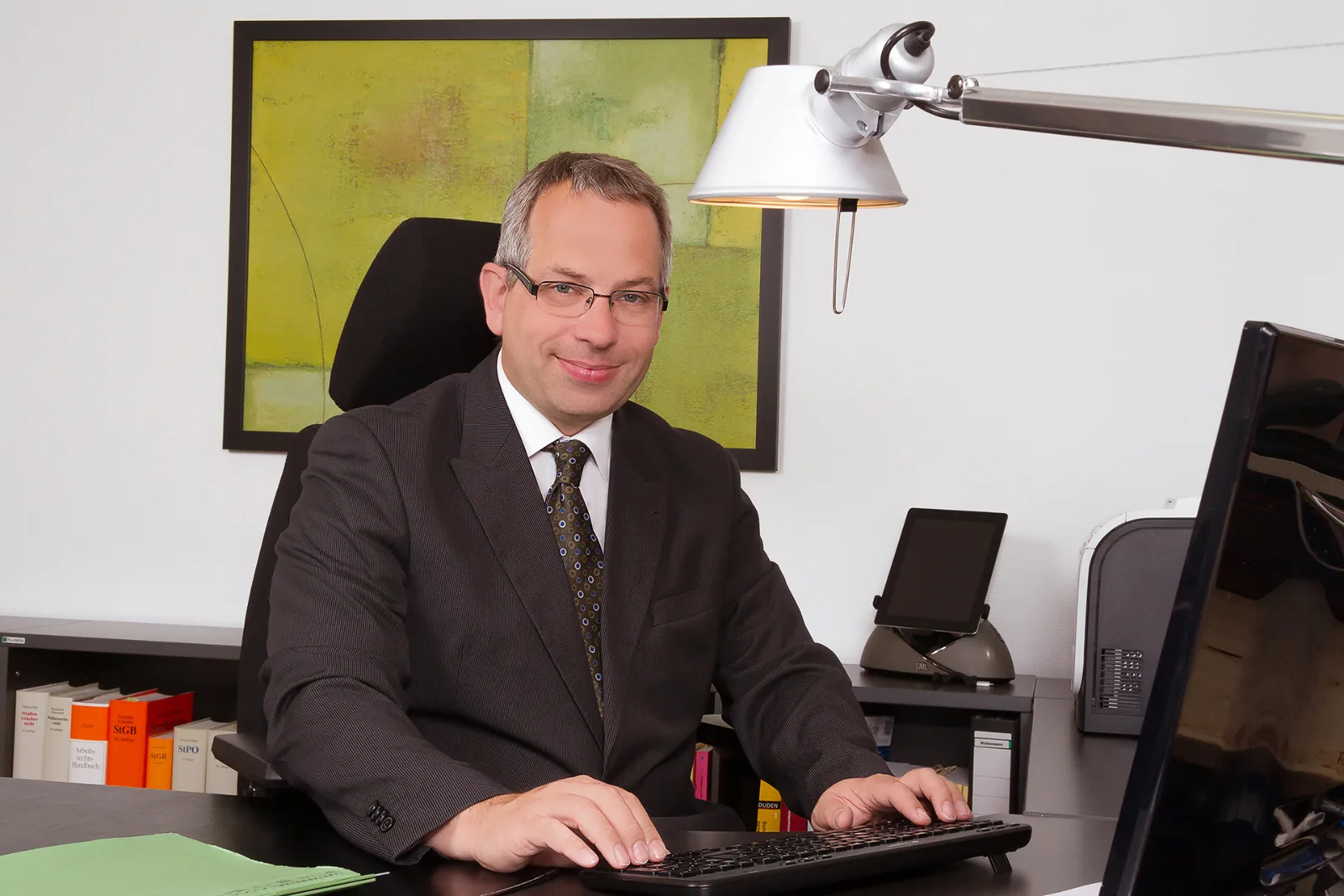 Businessman in a suit at his desk, with file spines and a modern painting in the background
