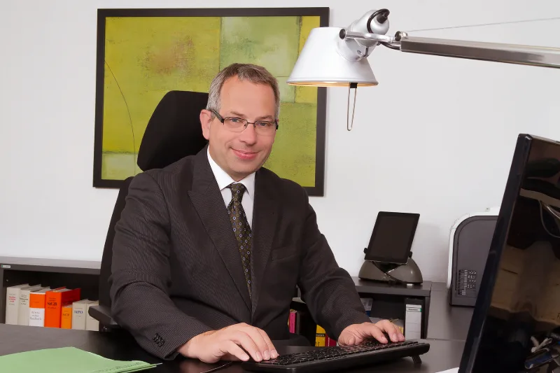 Businessman in a suit at his desk, with file spines and a modern painting in the background