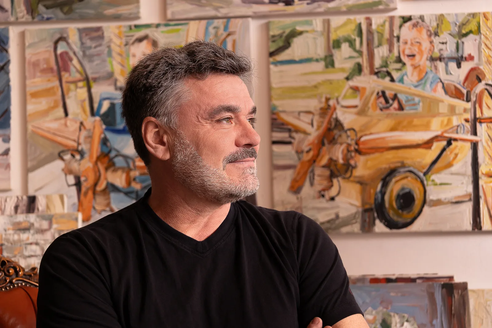 A man in a black T-shirt in front of several large oil paintings in his own studio