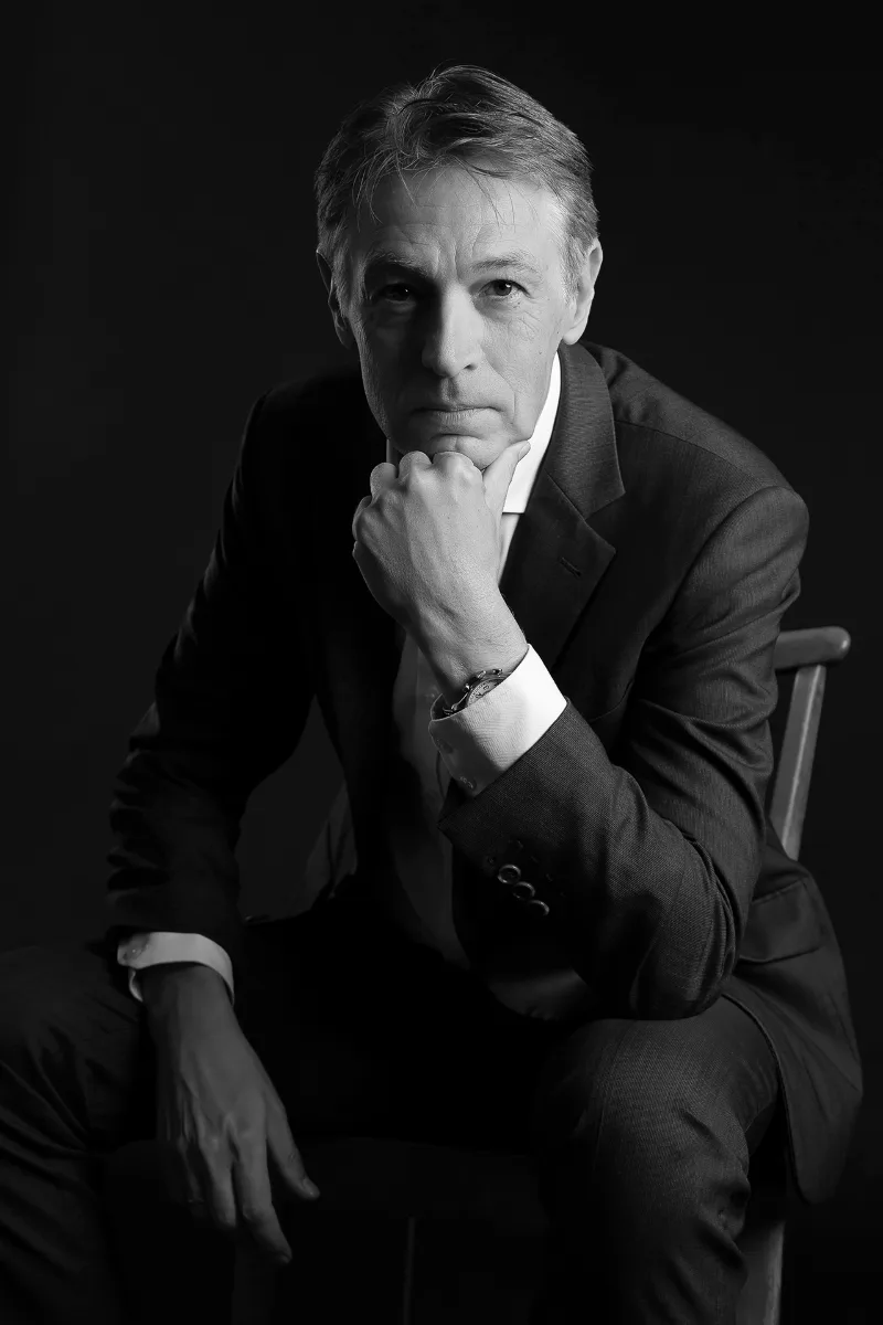 Black-and-white studio portrait: man in a suit on a chair, hand to chin, dark background