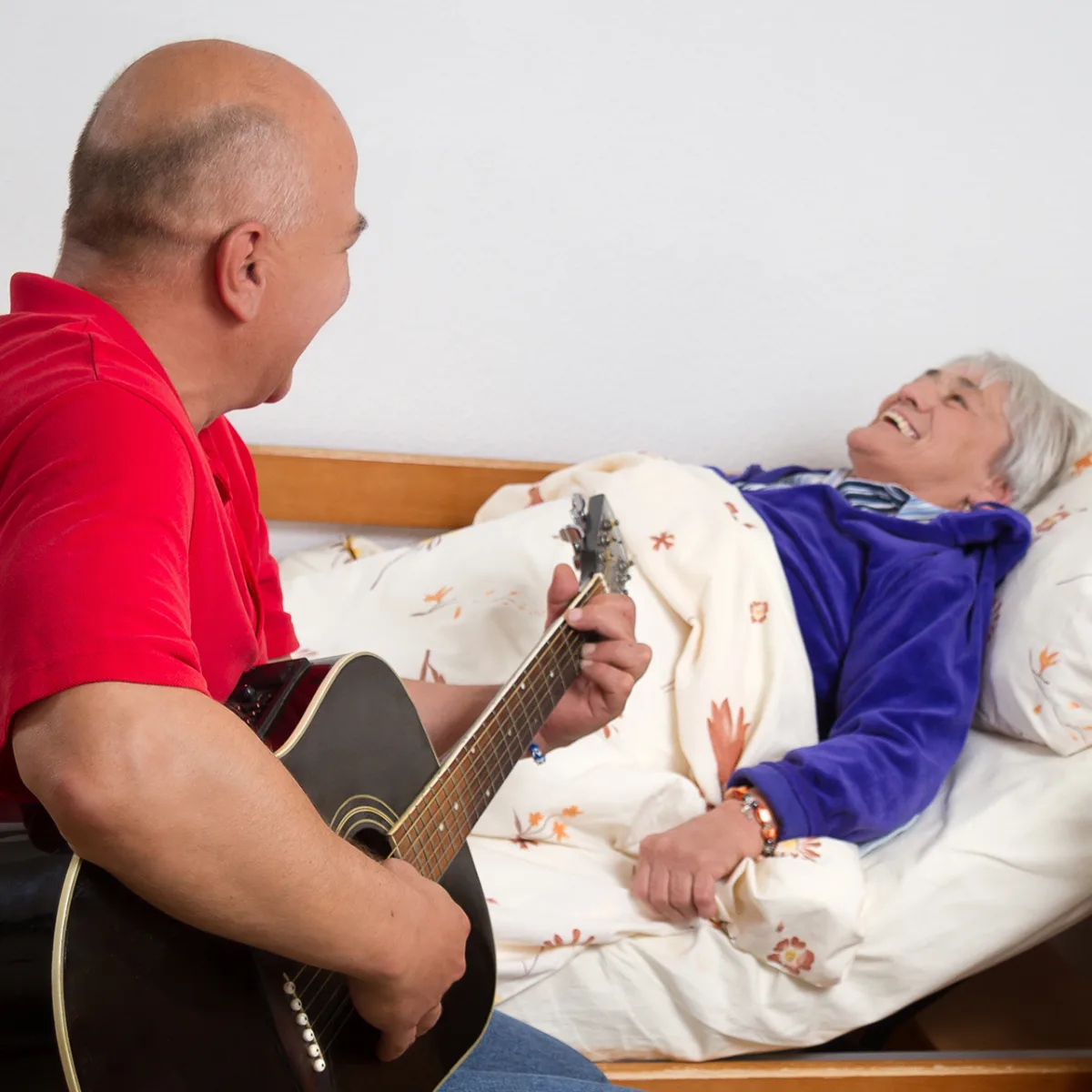 A carer in a red polo shirt plays acoustic guitar for a laughing patient in bed