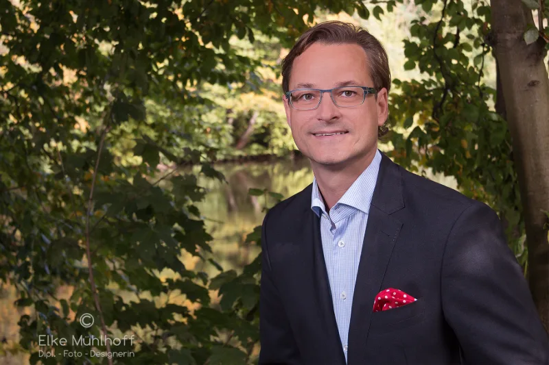 Geschäftsmann mit Brille und Einstecktuch vor herbstlichem Park mit Wasserspiegelung