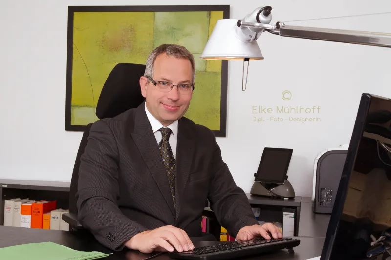 Anwalt mit Brille am Schreibtisch, im Hintergrund Aktenordner mit Gesetzestexten und modernes Kunstbild