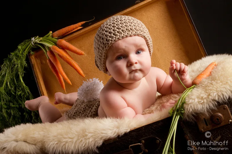 Baby mit gestrickter Hasenmütze sitzt in einem Koffer voller frischer Karotten