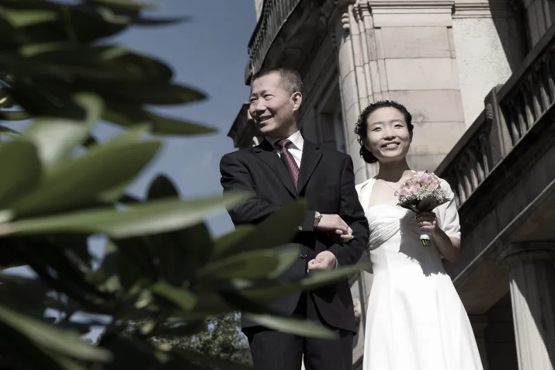 Bride and groom arm in arm in front of classical sandstone architecture, with a green hedge in the foreground