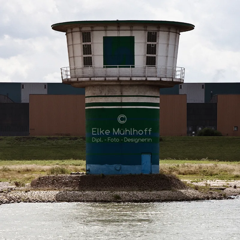 Bauchige Wachhütte auf einem grün-blauen Sockel am Rheinufer, im Hintergrund Industriehallen
