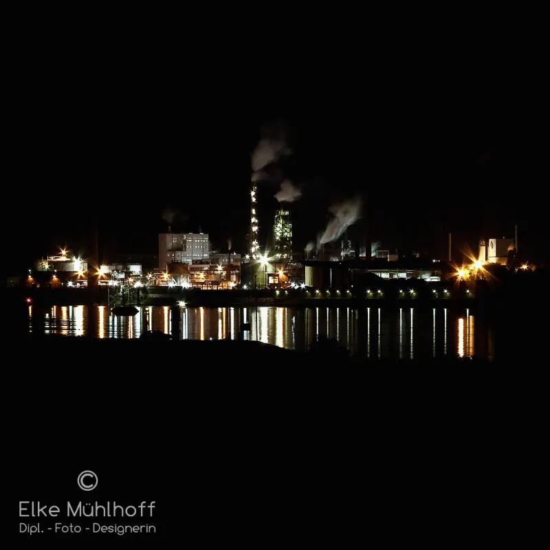 Beleuchtete Industrieanlage am Wasser in der blauen Stunde, Lichter spiegeln sich im Fluss