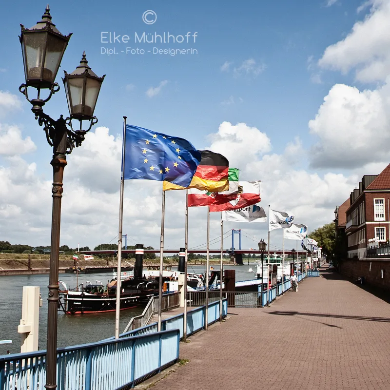 Sonnige Rheinpromenade mit reihenweise wehenden Flaggen, alter Schmuckstraßenlaterne und Ausflugsschiff
