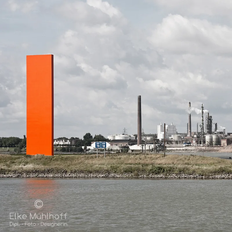 Großes orangenes Stahlmonument am Rheinufer, im Hintergrund die Türme eines Chemiewerks
