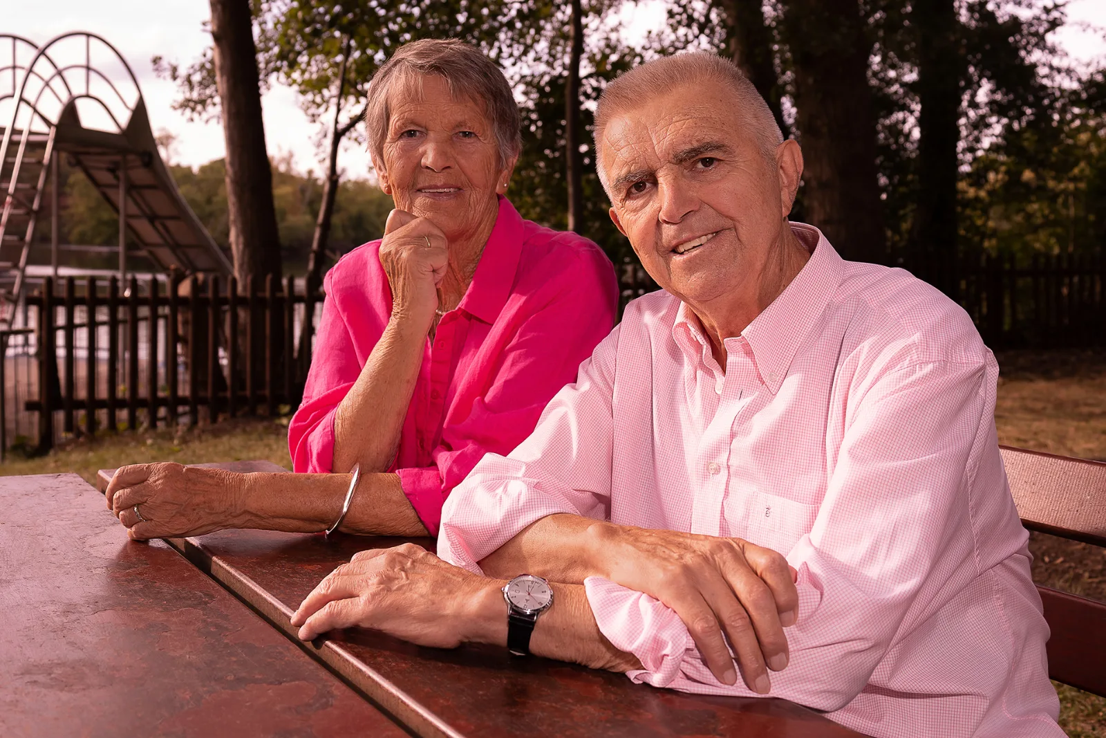 An older couple in pink and rose at a wooden table in the park, a lake in the background