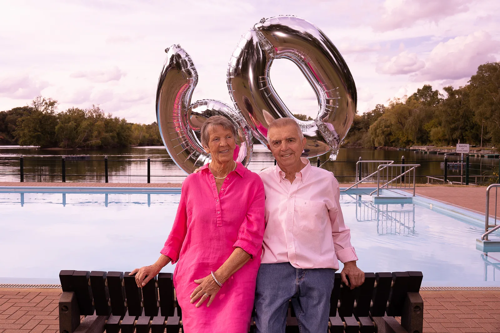 A couple in pink and rose on a wooden bench at the open-air pool, with silver helium balloons spelling 60 behind them