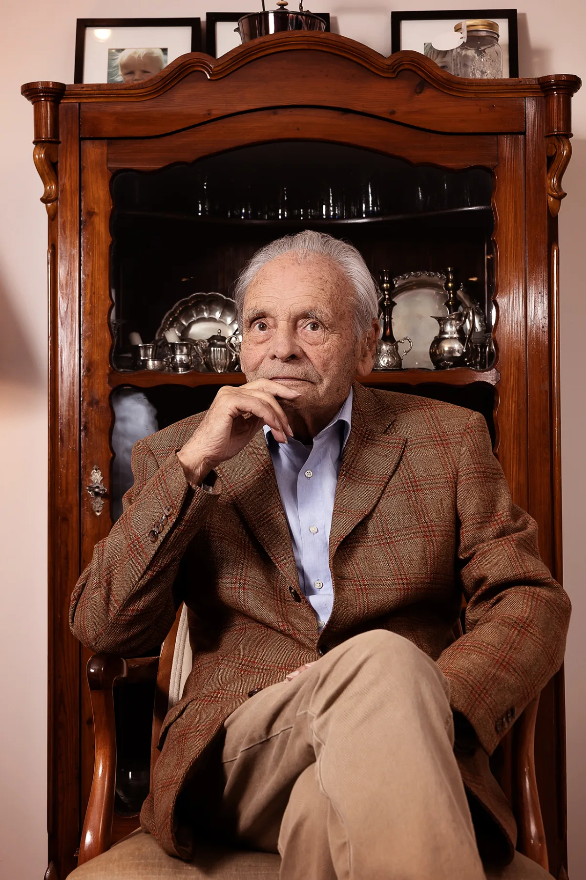 An older gentleman in a checked tweed jacket sitting thoughtfully in front of an antique display cabinet with silverware